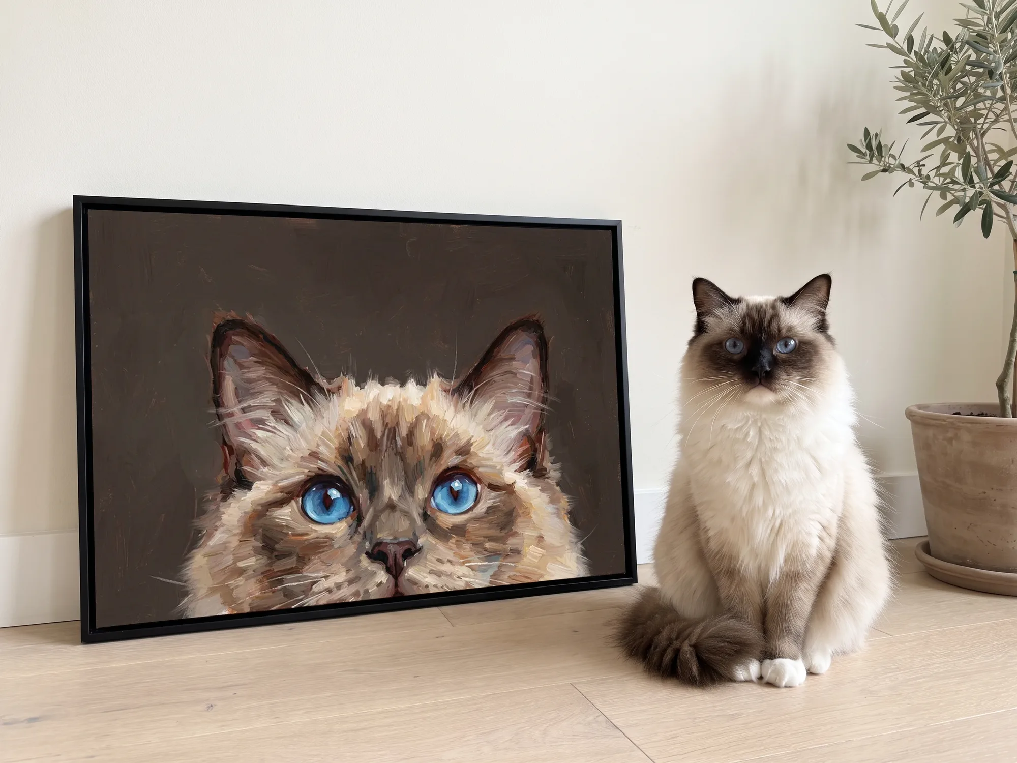 A ragdoll cat with blue eyes sitting on a wood floor beside a black-framed oil painting of the same cat, its head rising from the bottom edge of a dark canvas