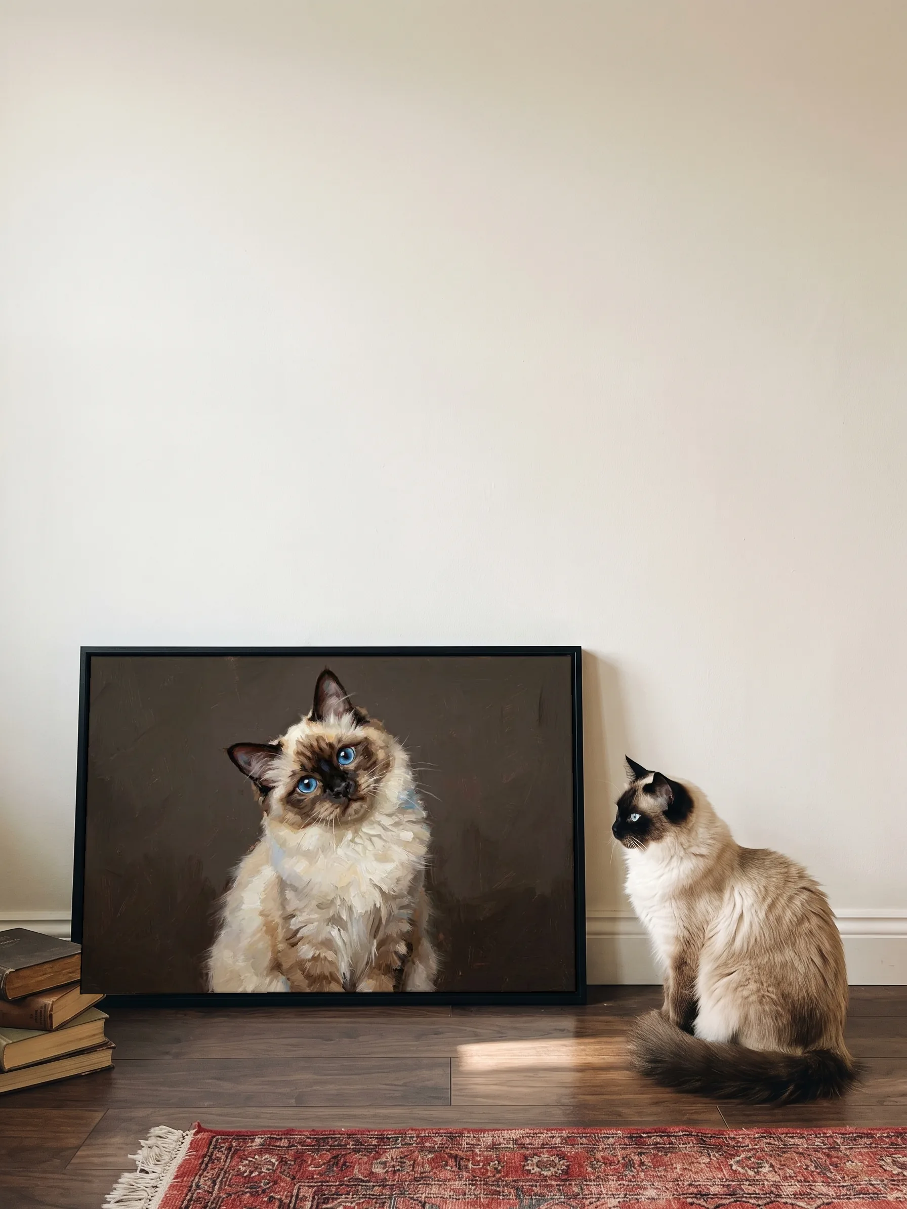 A ragdoll cat sitting in profile beside its own oil painting: a canvas in a slim black frame leaning on a wall, sun on the dark floorboards
