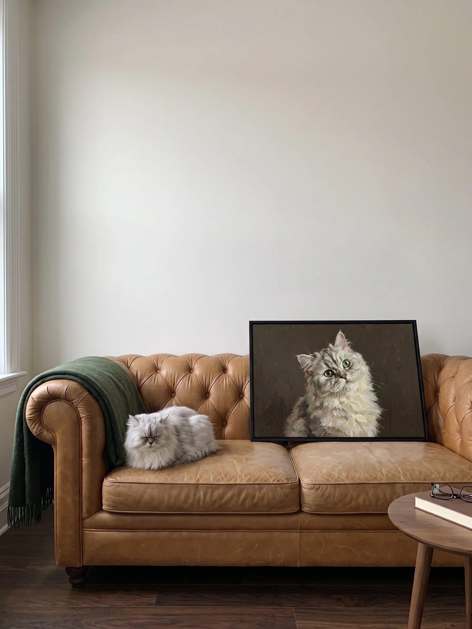 A silver Persian cat dozing on a tan leather sofa beside its own oil painting: a canvas in a slim black frame