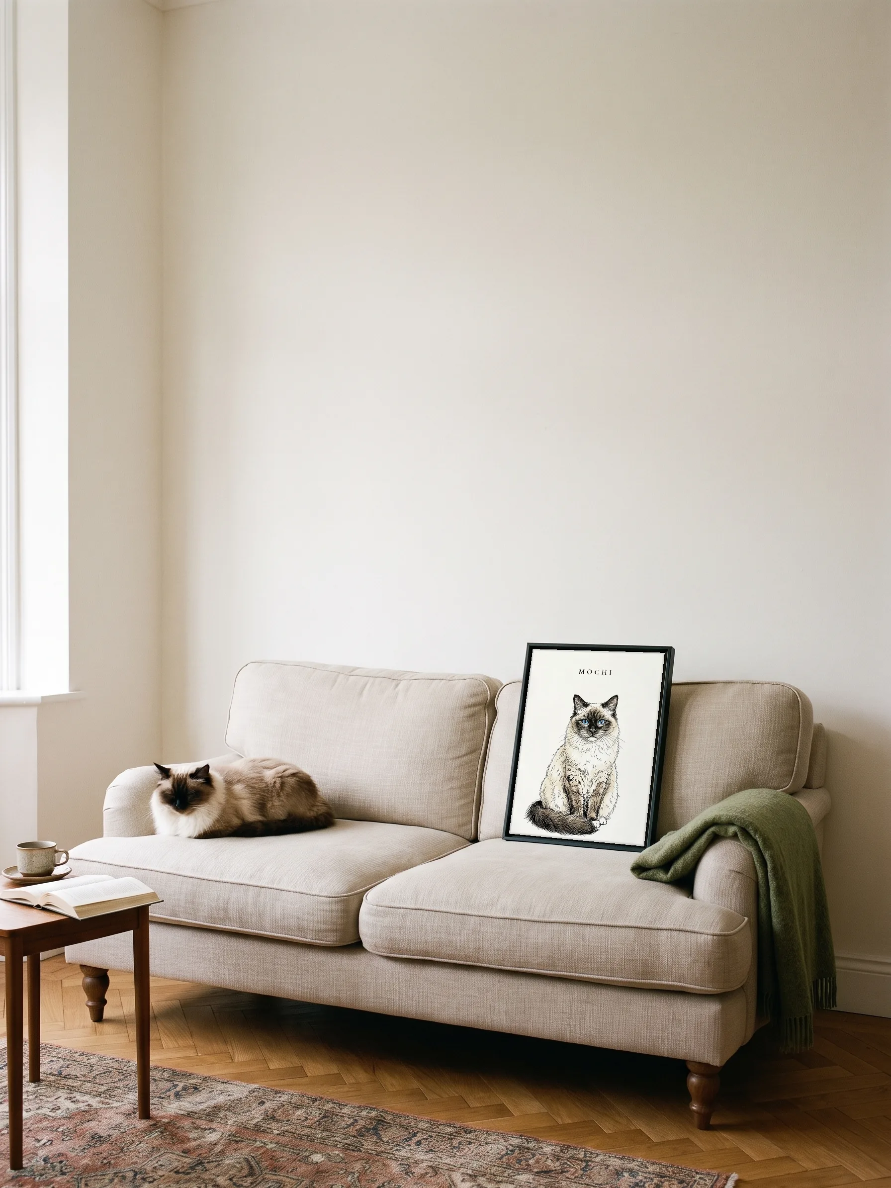 A ragdoll cat dozing on a linen sofa beside its own ink portrait: a canvas in a slim black frame