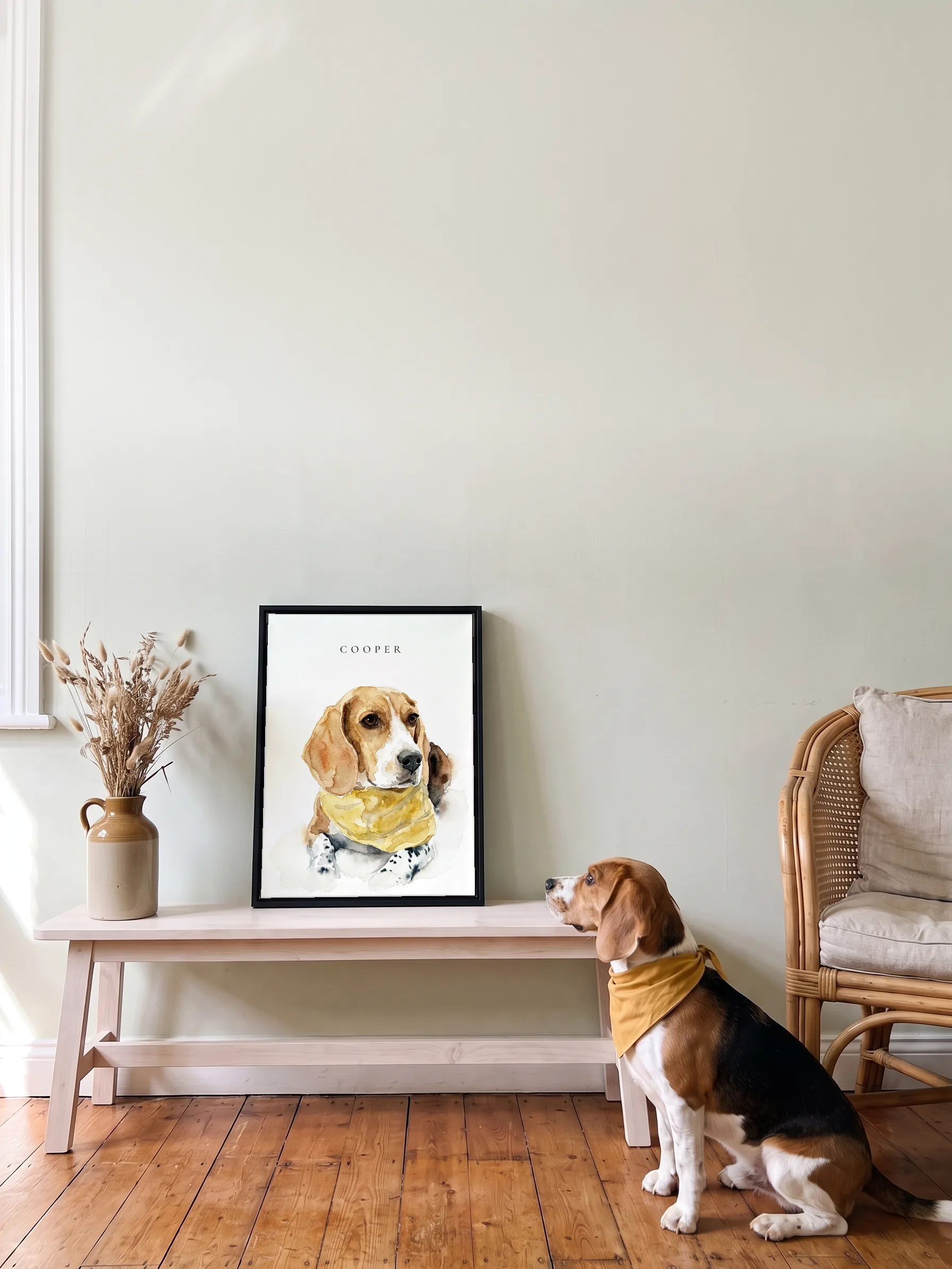 A beagle in a yellow bandana sitting on wooden floorboards, seen from behind, looking up at its own watercolor portrait: a framed canvas on a low pine bench
