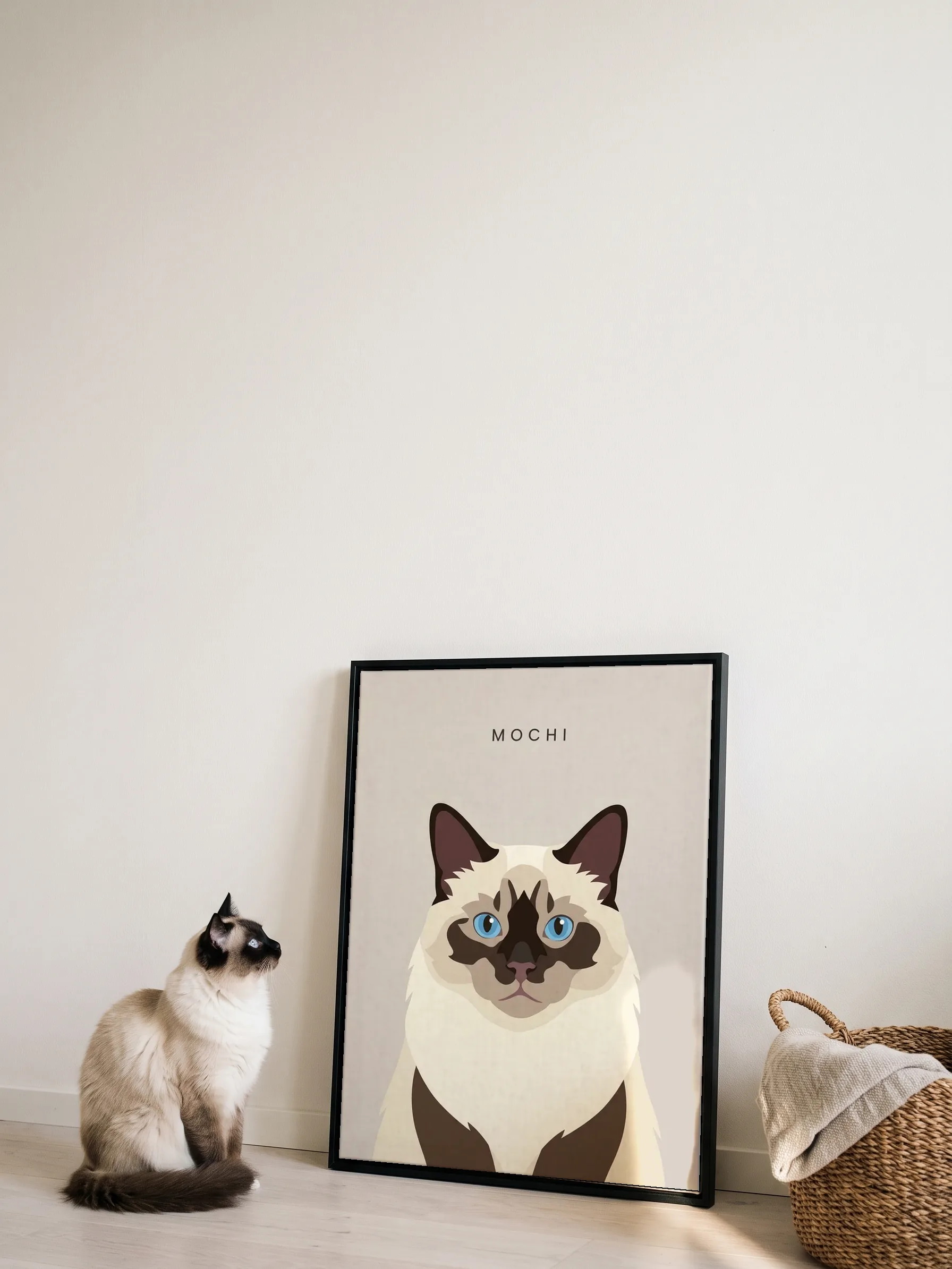 A ragdoll cat sitting in profile beside its own minimalist portrait: a canvas in a slim black frame leaning on a white wall, sun on its corner