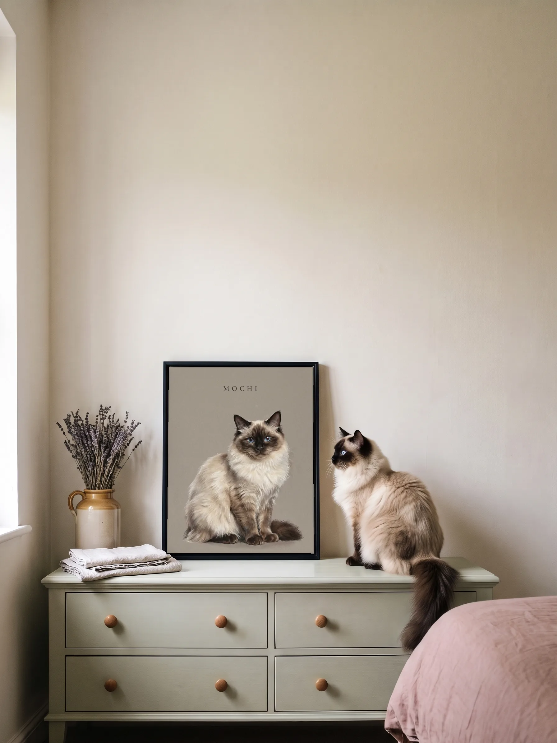 A ragdoll cat sitting on a sage green chest of drawers, seen from behind, beside its own pastel portrait: a framed canvas leaning on the wall