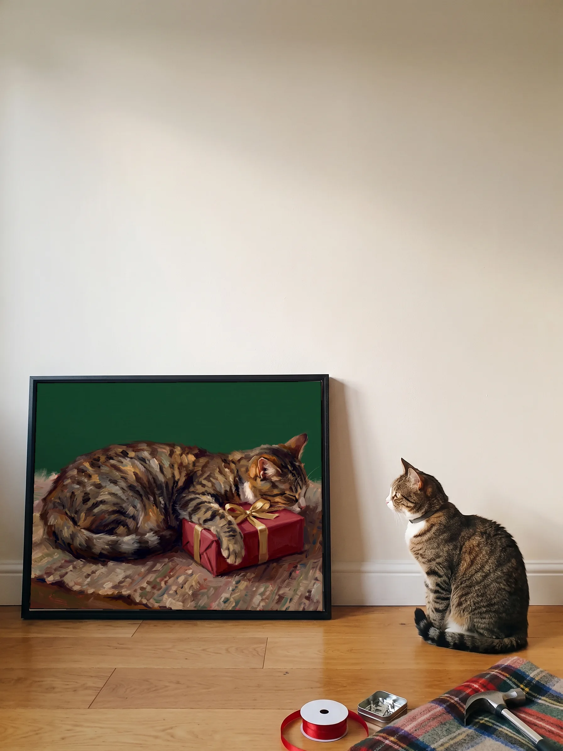 A tabby cat sitting beside its own oil painting: a wide canvas in a slim black frame leaning on a wall, a spool of red ribbon on the floor